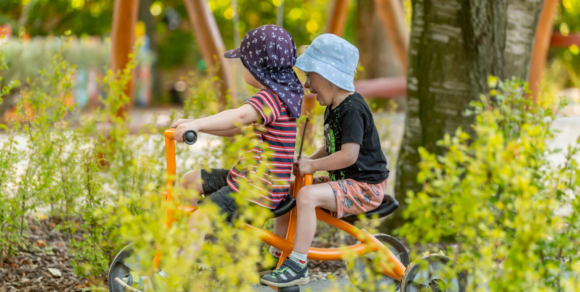 Zwei Kinder mit Sonnenhüten fahren im Schatten Dreirad