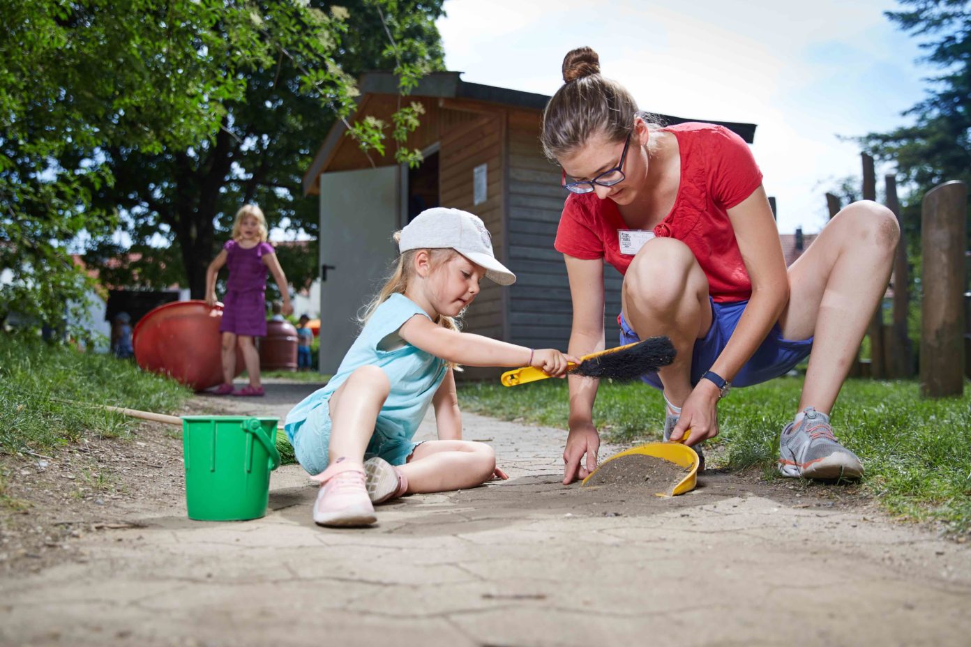 Ein kleines Mädchen hilft ihrer Erzieherin beim Fegen eines Gartenweges der Kita.
