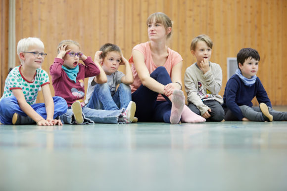 Eine Erzieherin und fünf Kinder sitzen in der Turnhalle der Kita.