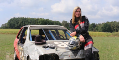 Das Foto zeigt eine junge Frau, die in einem Rennanzug gekleidet und gestützt auf einen Remmhalm auf der Motorhaube eines ramponierten Autos sitzt. Davor steht ein kleines, rotes Rutschauto (Bobbycar).