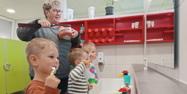 Drei Kinder stehen vor einem Spiegel und putzen sich die Zähne. Hinter ihnen steht eine Erzieherin und putzt einem großen Gebiss die Zähne.