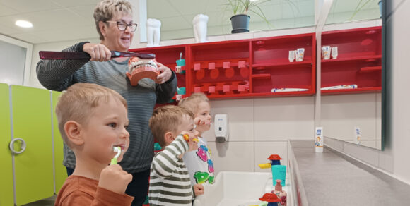 Drei Kinder stehen vor einem Spiegel und putzen sich die Zähne. Hinter ihnen steht eine Erzieherin und putzt einem großen Gebiss die Zähne.